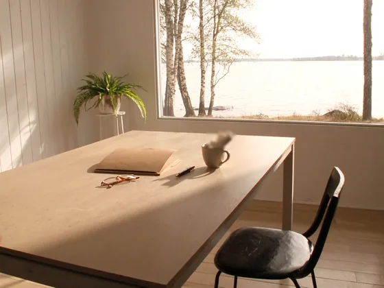 A pale wooden table by a large window onto a lake, with a closed folder, reading glasses, a pen and a cup, a fern on a stand behind